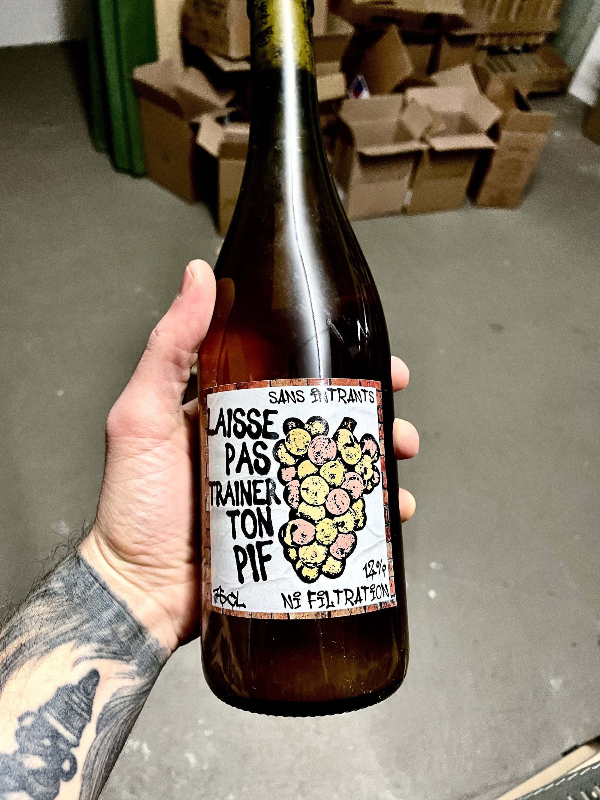 Hand holding a bottle with a label in an indoor setting with cardboard boxes in the background