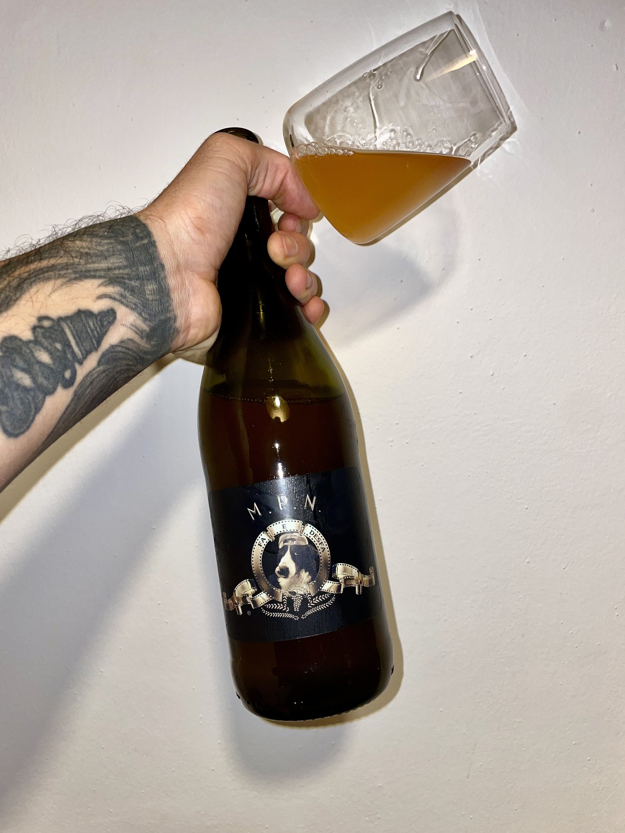 Hand pouring beer from a bottle labeled 'M.P.N.' into a glass on a white background