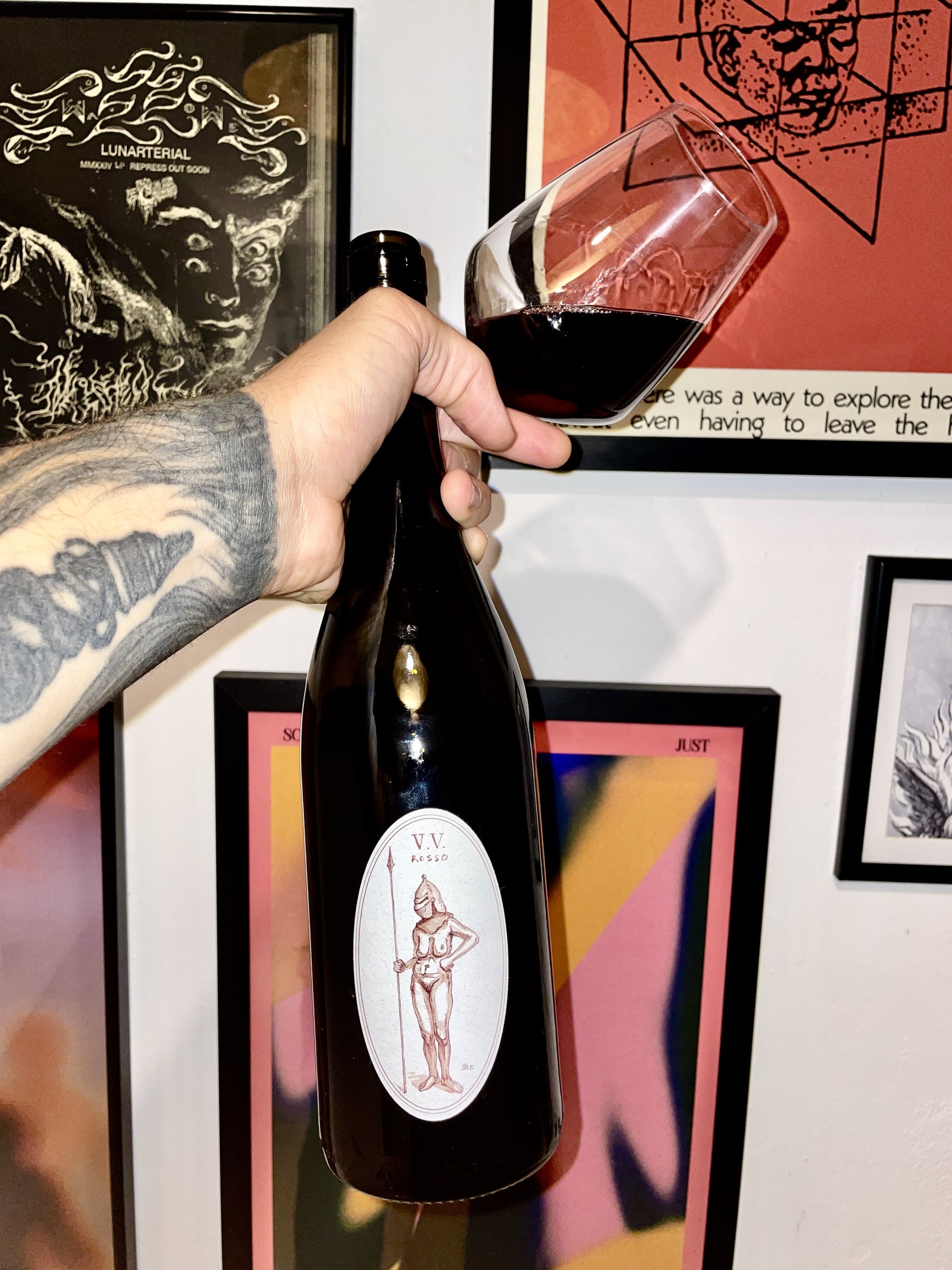 Person holding a bottle of wine with a glass, in front of framed artwork on a wall.