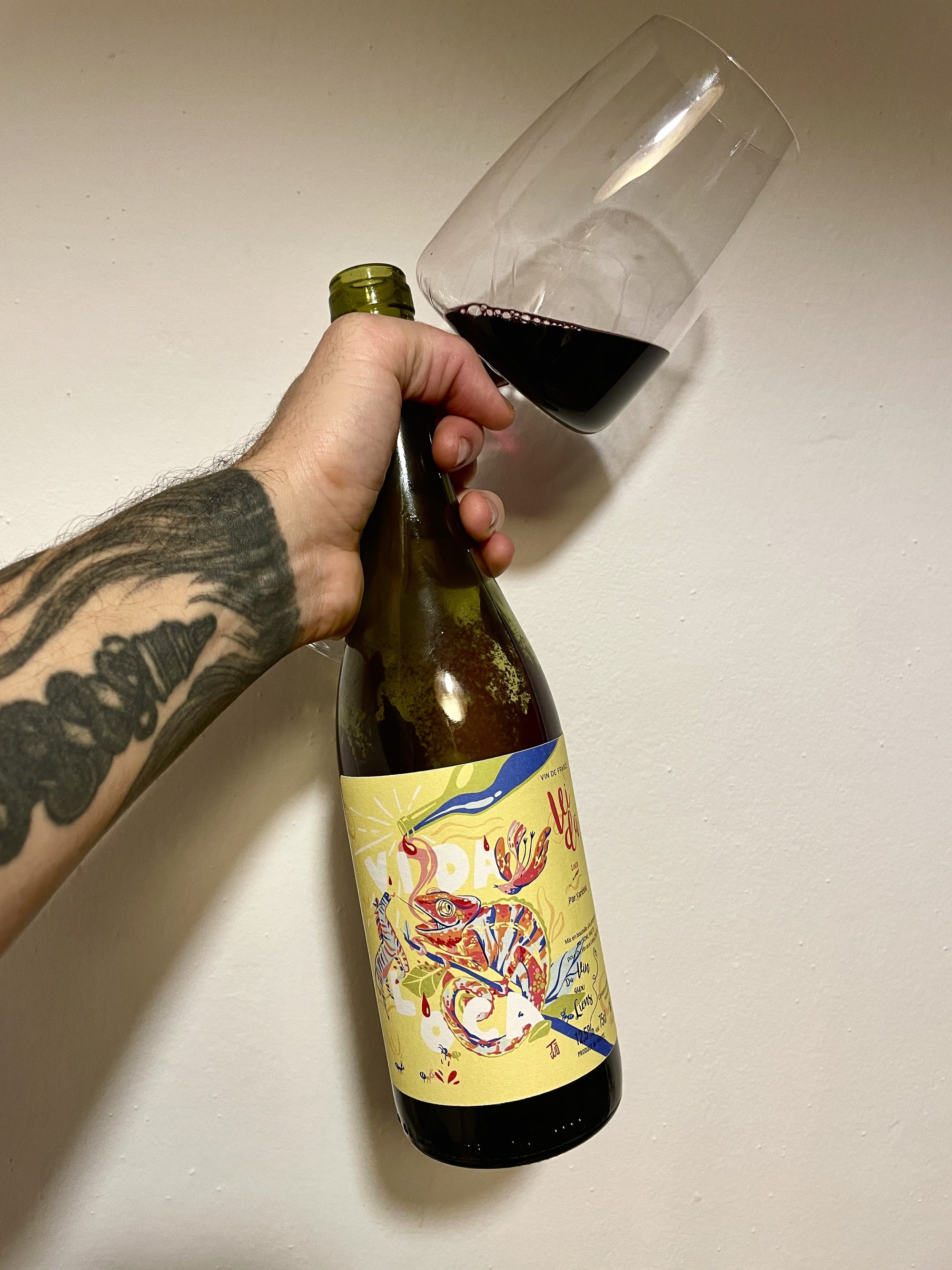 Person pouring red wine from a bottle with a colorful label into a glass on a plain background