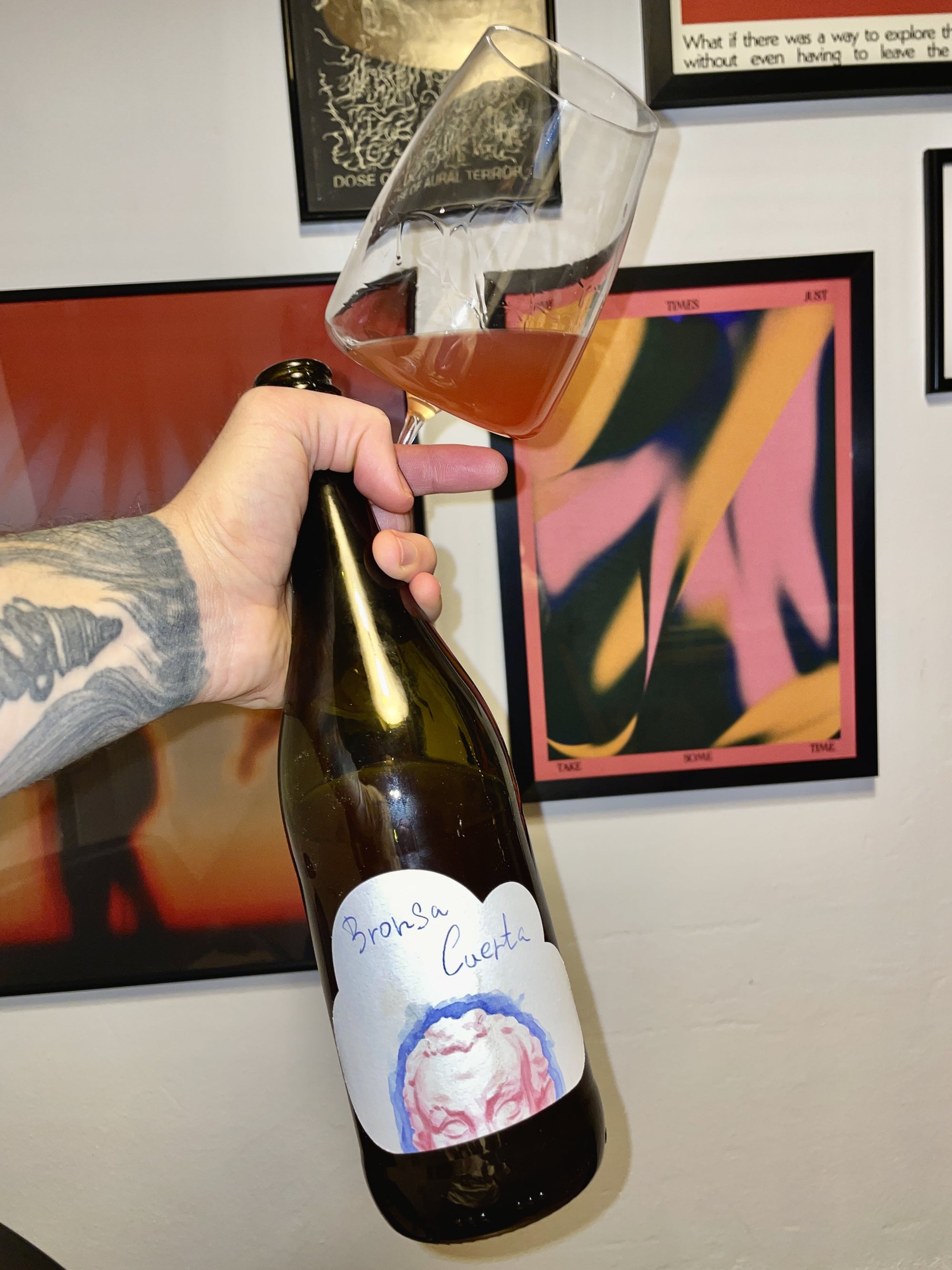 Hand holding a wine glass with a bottle labeled 'Bronsa Cuerta' against a colorful abstract painting background.