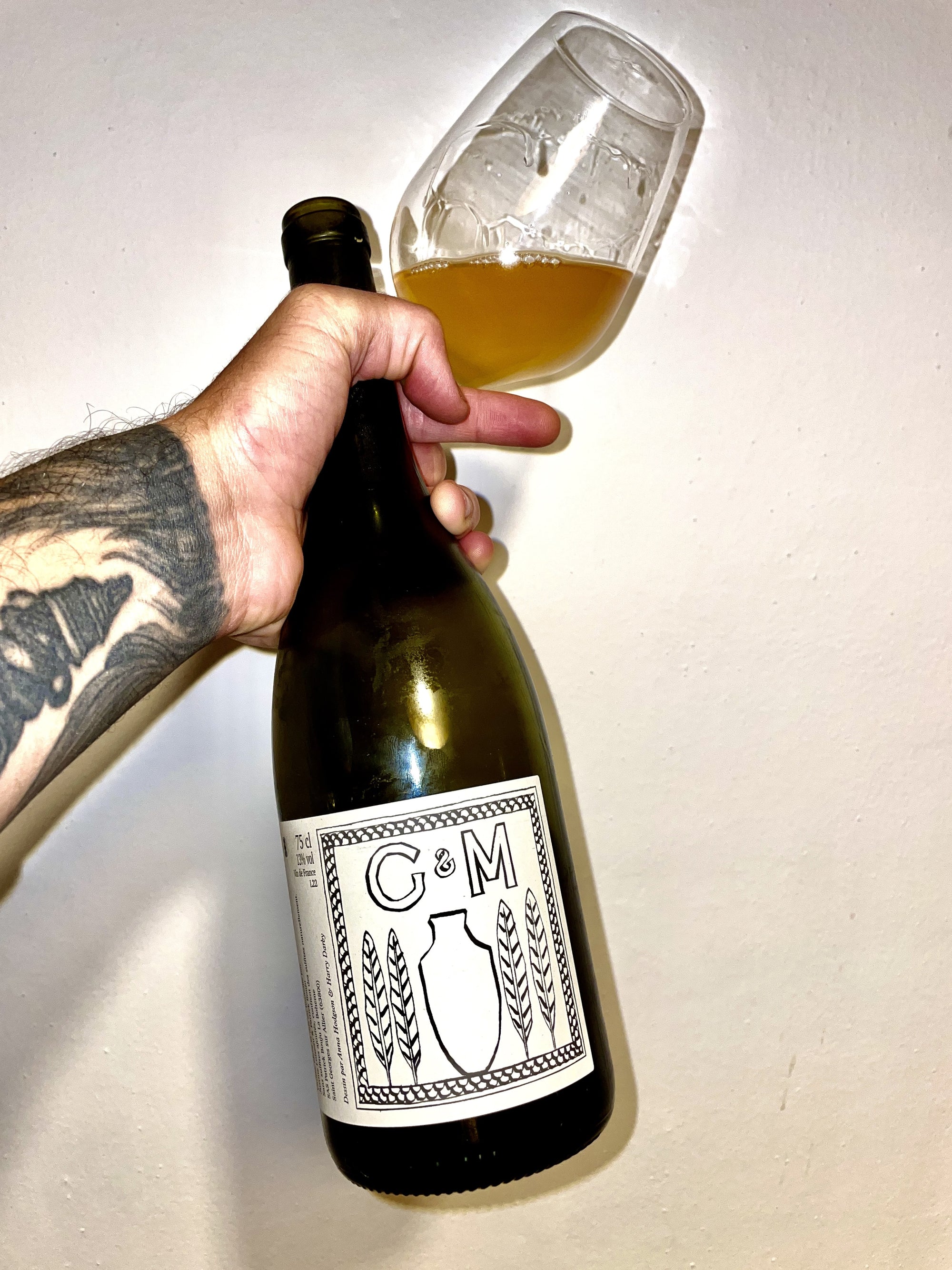 Hand holding a bottle of wine with a glass filled with orange wine, against a plain background