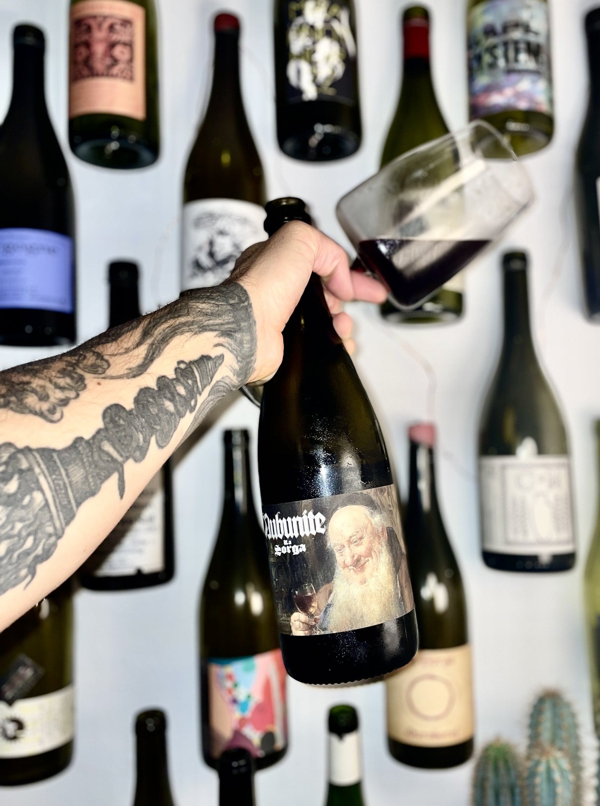 Person pouring wine from a bottle labeled 'Aubunite' into a glass against a backdrop of various wine bottles.