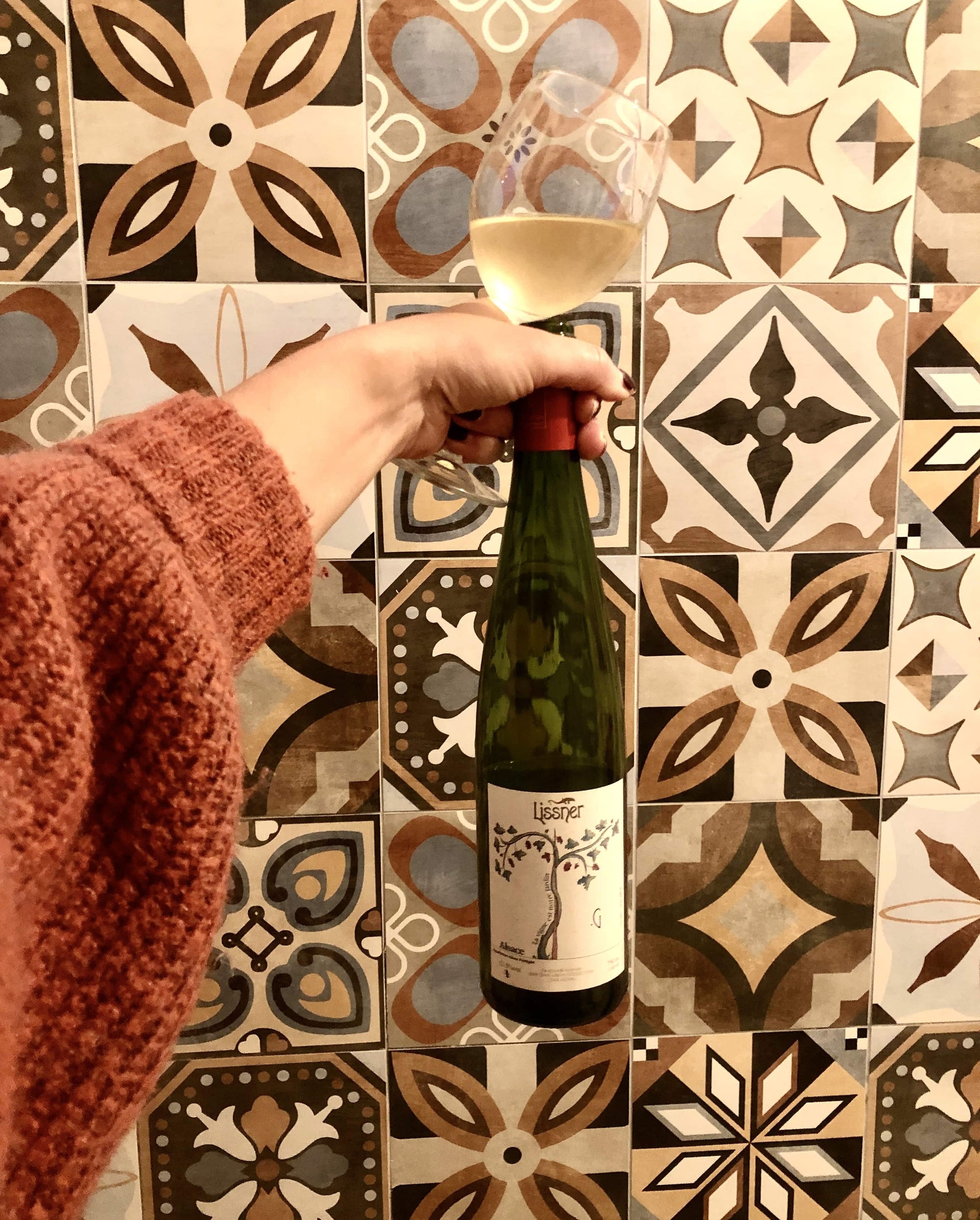 A bottle Of Edelzwicker White Wine and a glass of white wine in front of a colorful Tile Wall