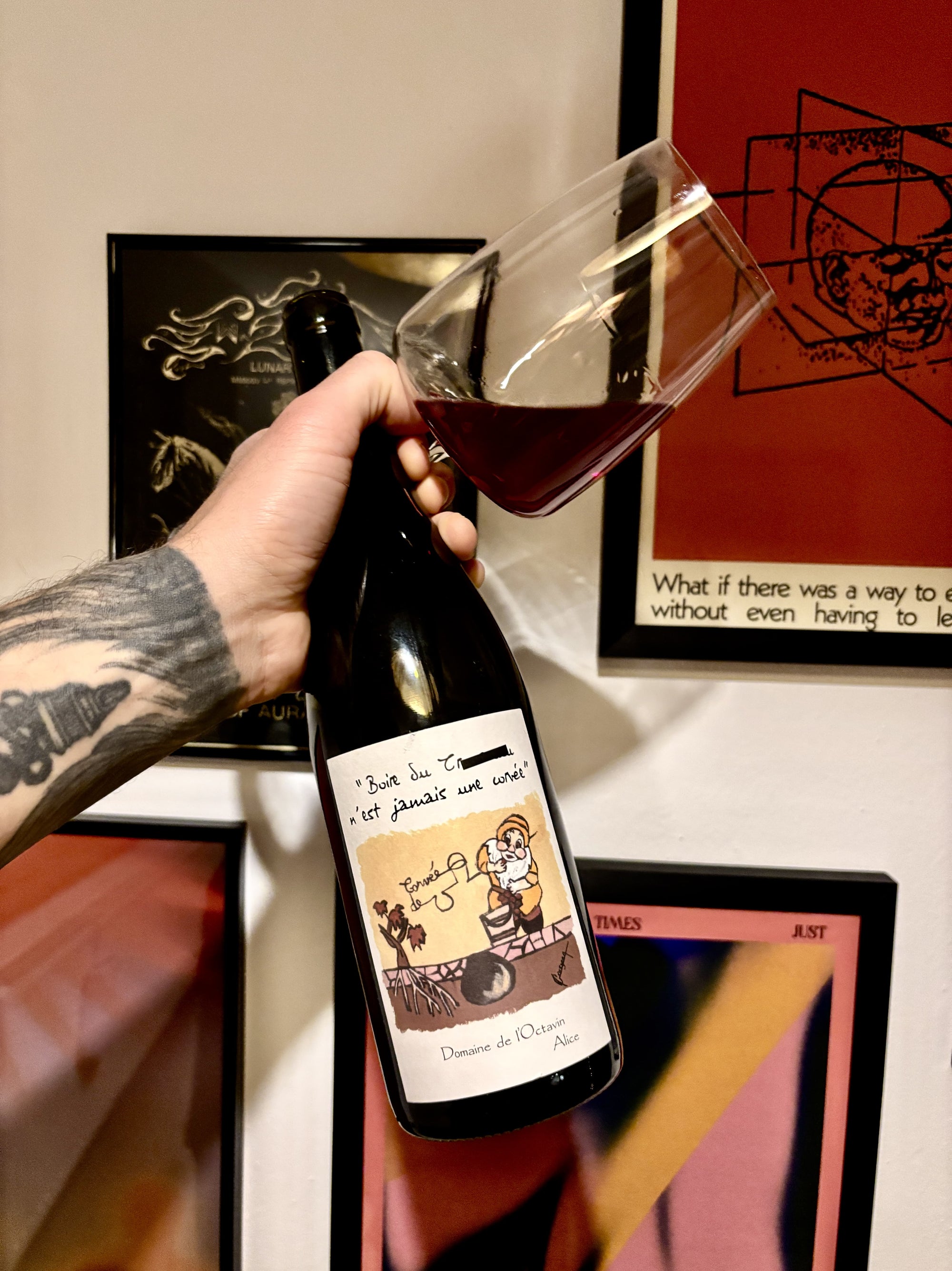 A bottle and glass of red wine held by hand in front of a wall with artwork
